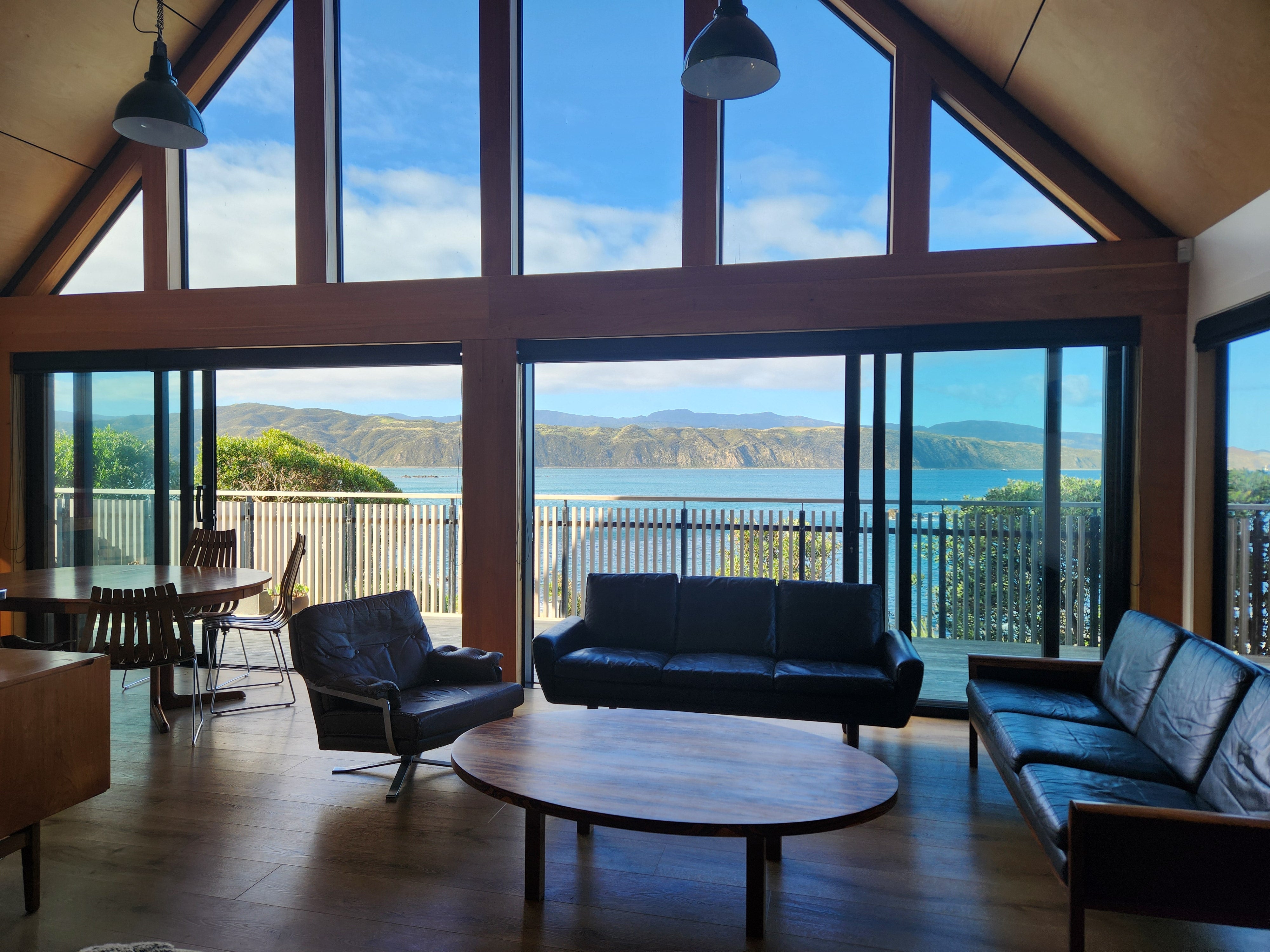 Pictorial image of a mid century living room with mid century furnishings. Set within a 1960's building with an A-frame roof. With a view to sea with mountains and blue sky.