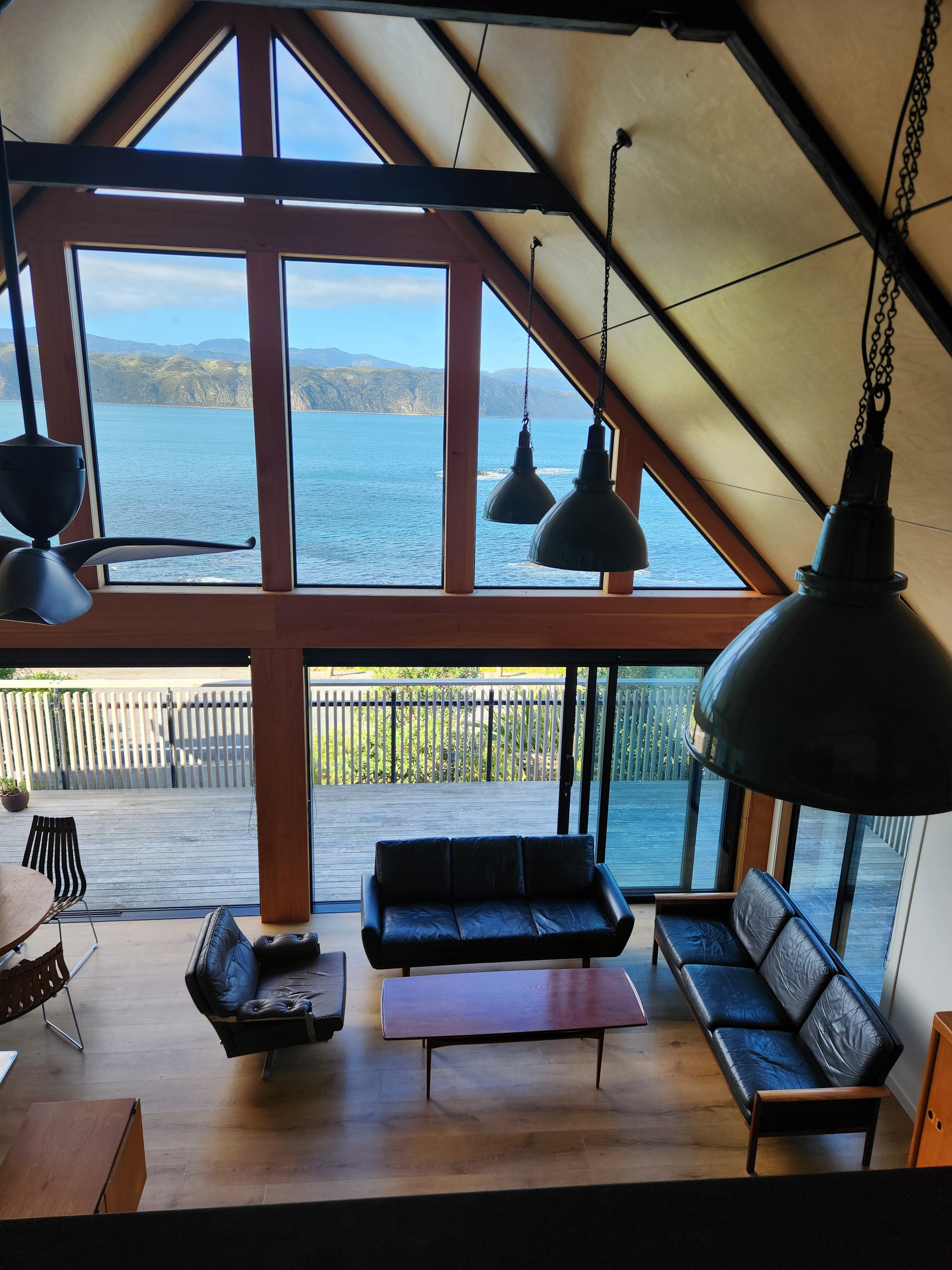 Pictorial image of a mid century living room with mid century furnishings and industrial lighting. Set within a 1960's building with an A-frame roof. With a view to sea with mountains and blue sky.