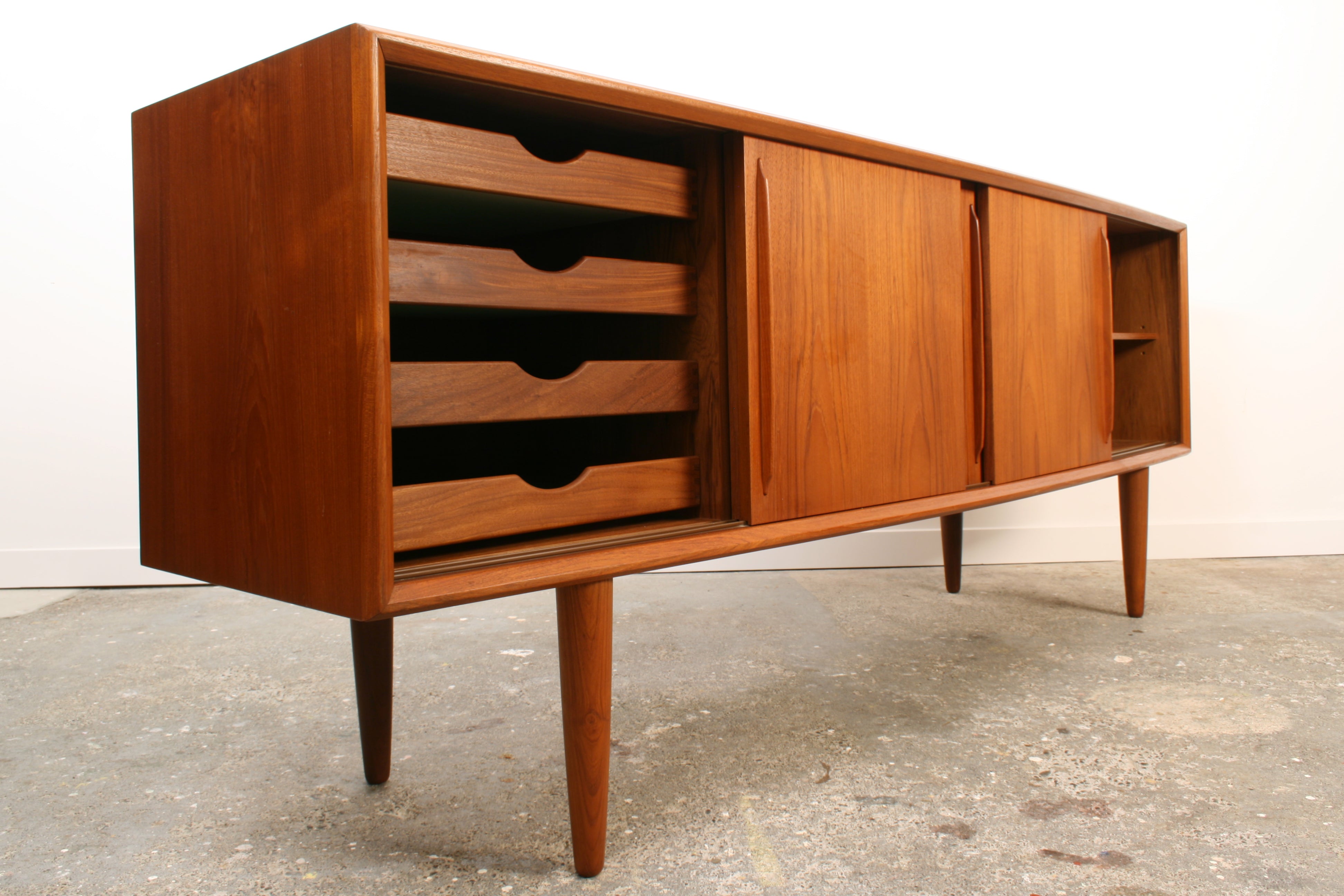 Danish Teak sideboard by H P Hansen. Shown at a slight angle from the left. It has four sliding doors. The outer two are open. The left revealing four sliding drawers and on the right an adjustable shelf.